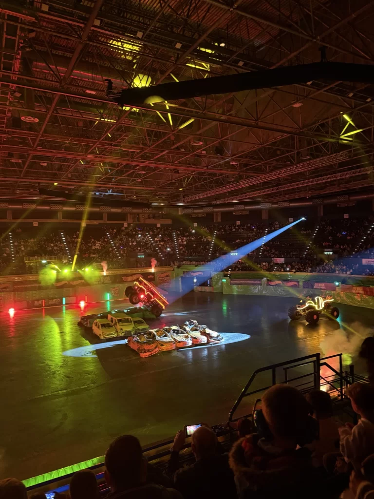 Indoor arena with monster trucks performing stunts, colorful lights shining, and spectators watching from the stands.