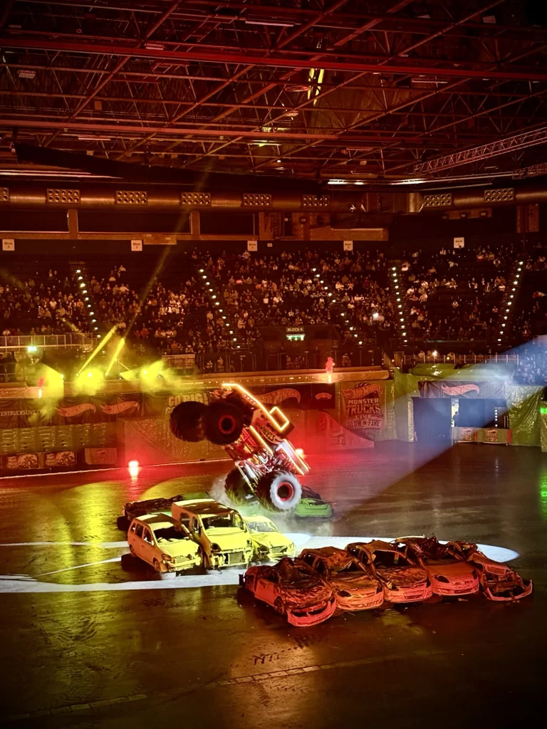 A monster truck performing a jump over crushed cars in an arena with a large audience and dramatic lighting.