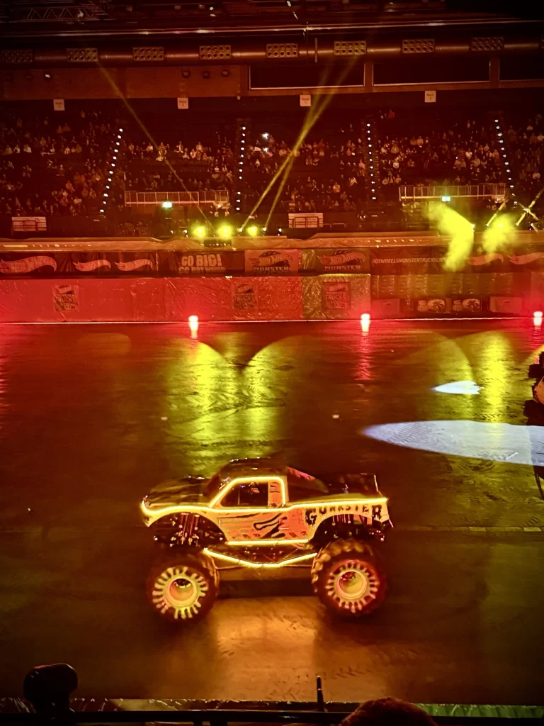 A monster truck with "Gunslinger" on its side drives in an indoor arena with colorful lights and a crowd in the background.
