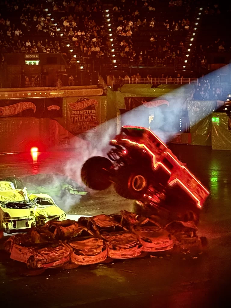 A monster truck with neon lights jumps over a pile of crushed cars in an indoor arena during a show, with an audience watching in the background.