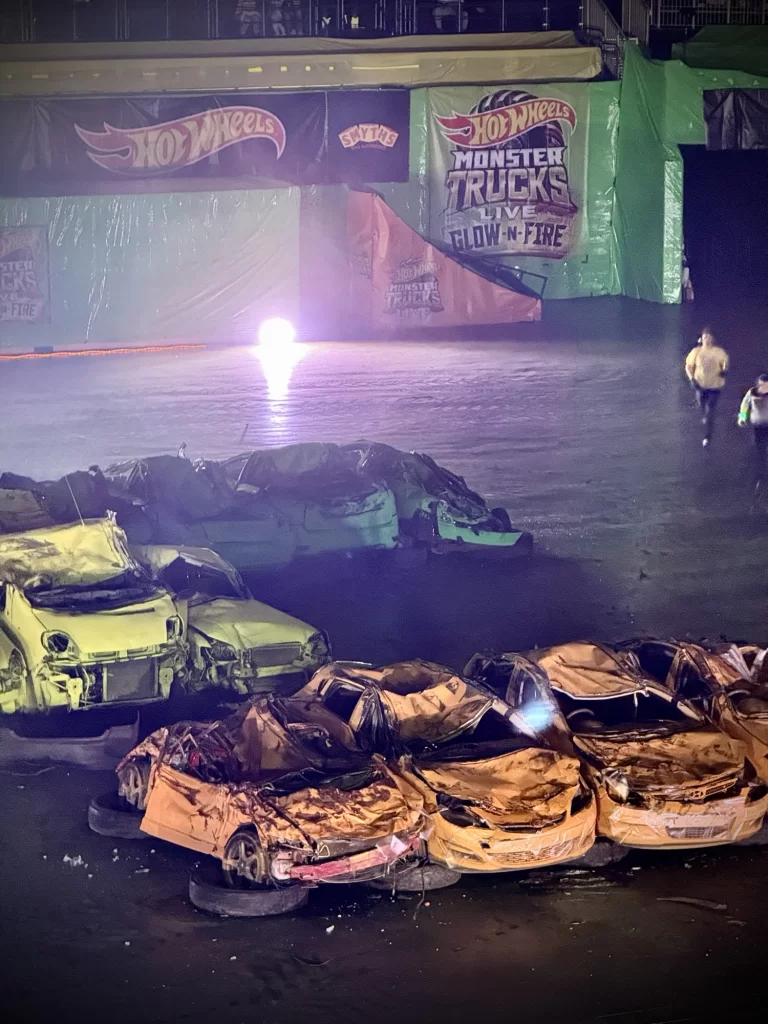 Crushed cars arranged in a circle on an arena floor, with a "Hot Wheels Monster Trucks" backdrop and two people walking nearby.