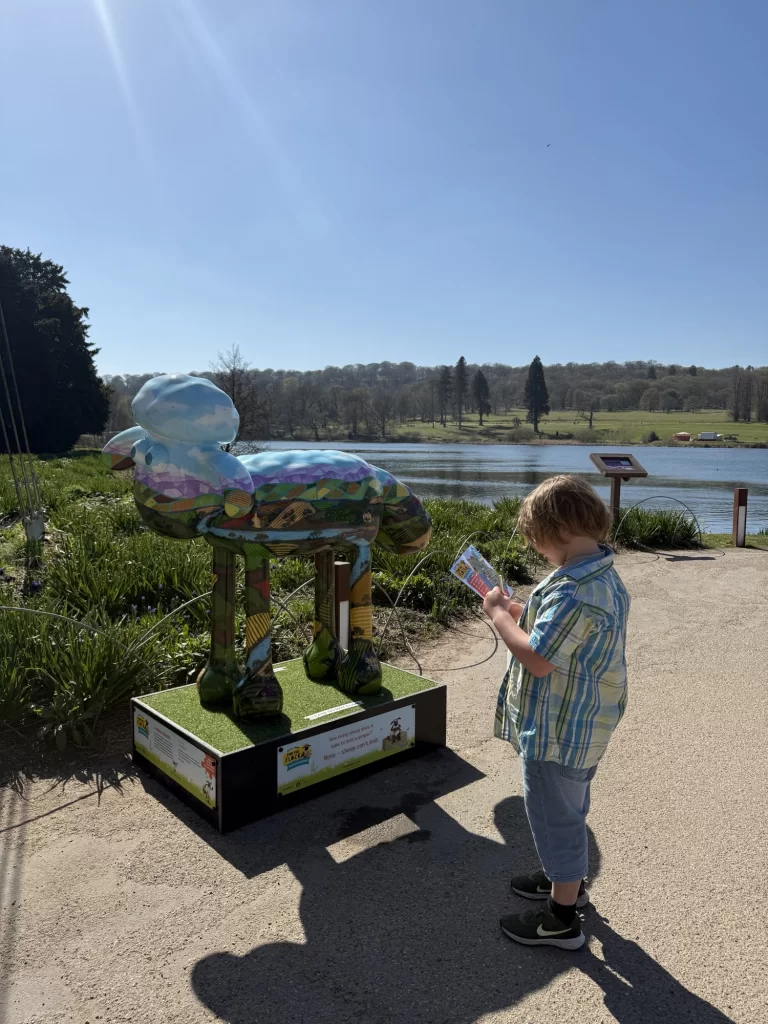 Child wearing a plaid shirt, looking at a booklet next to a colorful sheep statue, near a lake and grassy area under a clear sky.