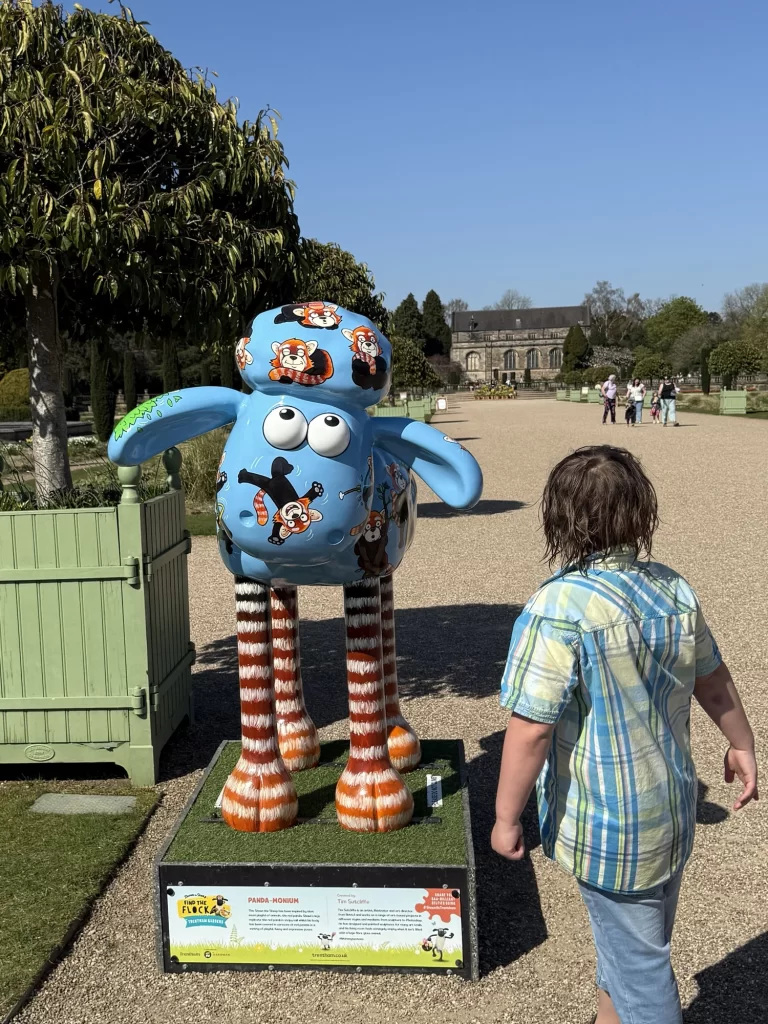 A child looks at a colorful Shaun the Sheep statue in a landscaped garden with a historic building in the background.