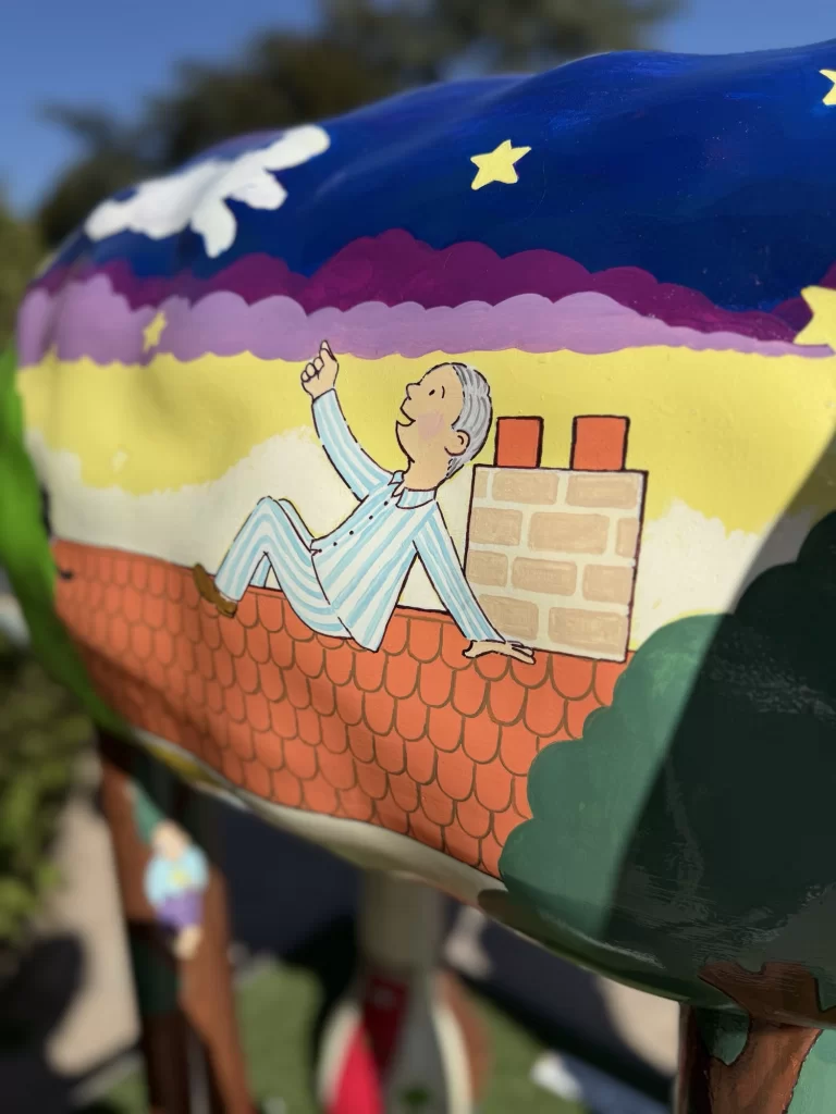 Colorful sculpture depicting a cartoon figure in pajamas lying on a rooftop, pointing at the night sky with stars and clouds.