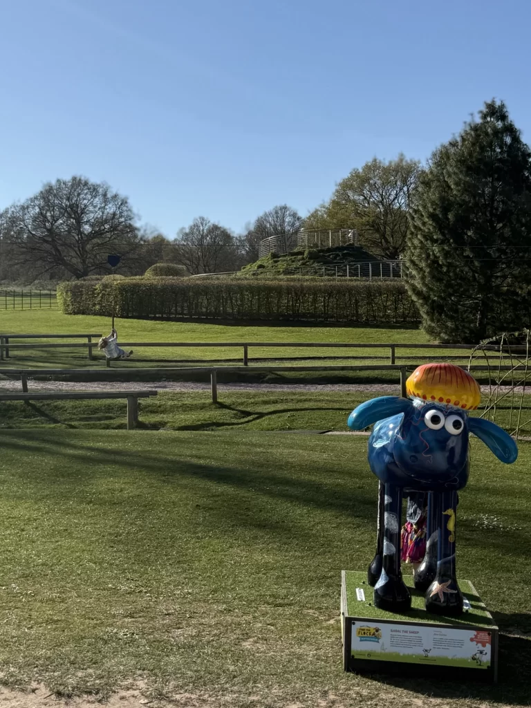 Colorful sheep sculpture on grass with trees and hedges in the background on a sunny day.