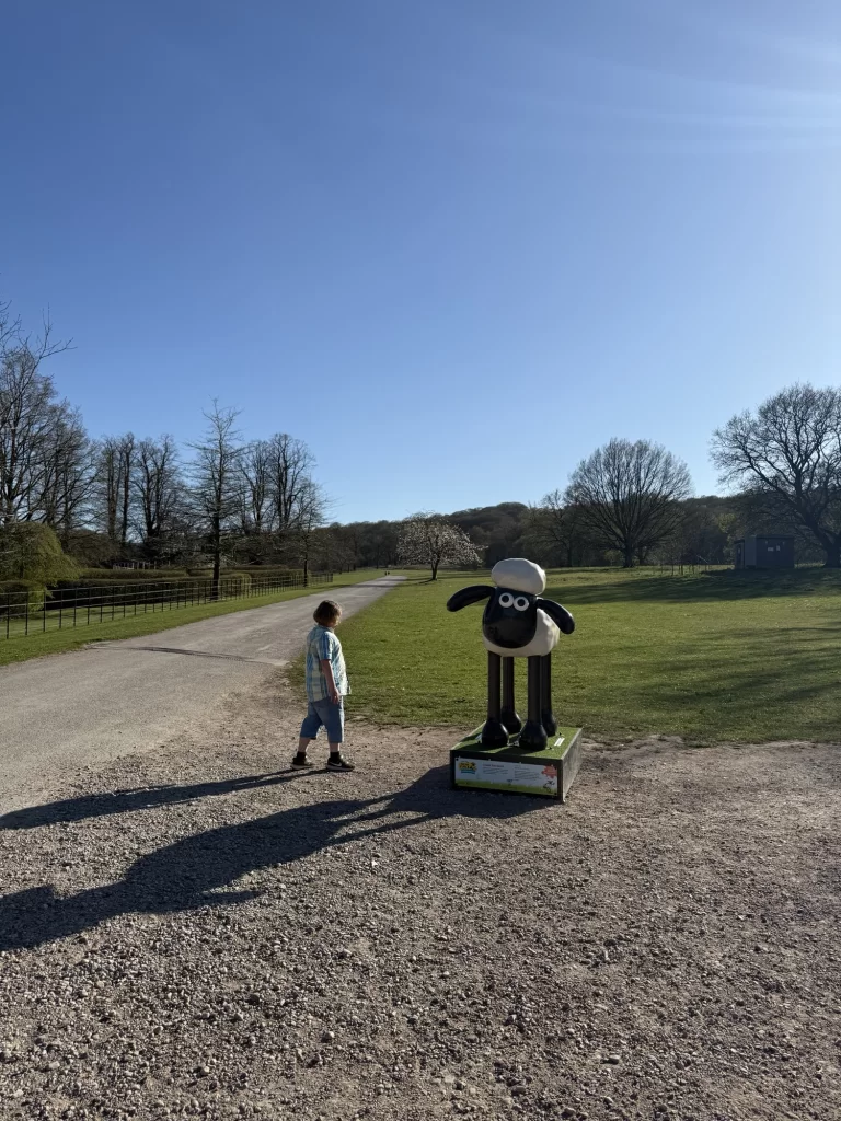 Child walking on gravel path toward a Shaun the Sheep statue in a park with grass, trees, and a clear sky in the background.