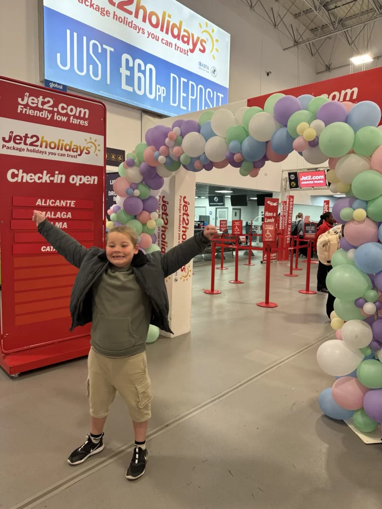 A young boy stands smiling with arms raised under a pastel balloon arch at a Jet2 check-in area inside an airport terminal.