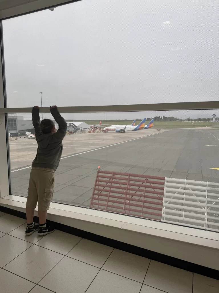 A person in shorts and a hoodie looks out an airport window at airplanes parked on the runway under an overcast sky.
