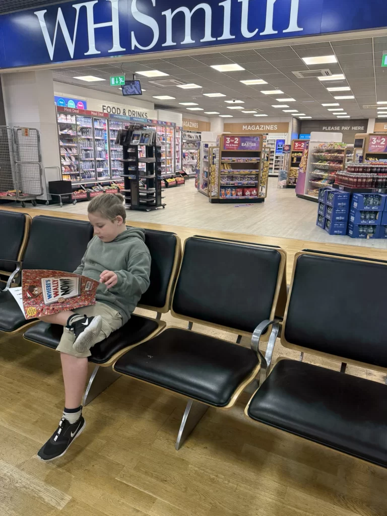 A young boy sits on a bench reading a magazine in front of a WHSmith store inside a shopping area.