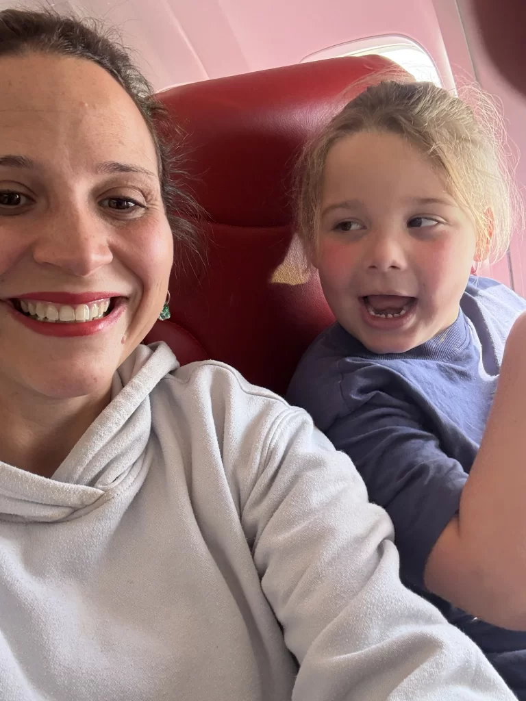 An adult and a child sit next to each other on red airplane seats, smiling and looking at each other.