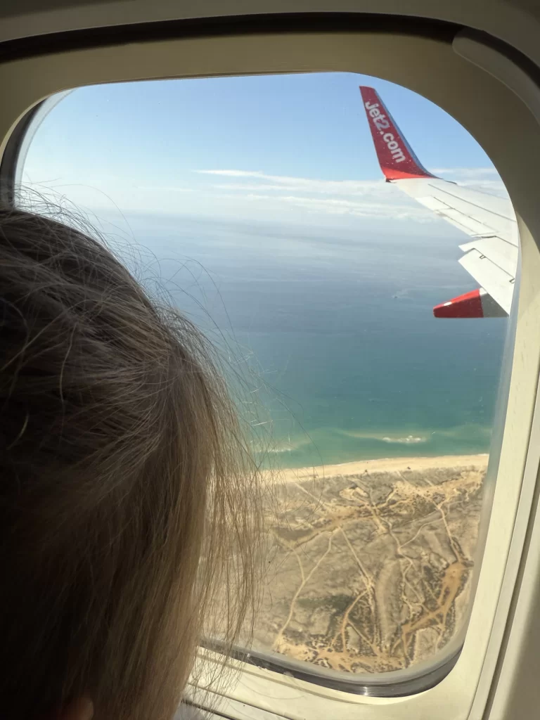 A person looks out an airplane window at a coastline and ocean below, with part of the plane's wing and a "jet2.com" logo visible.