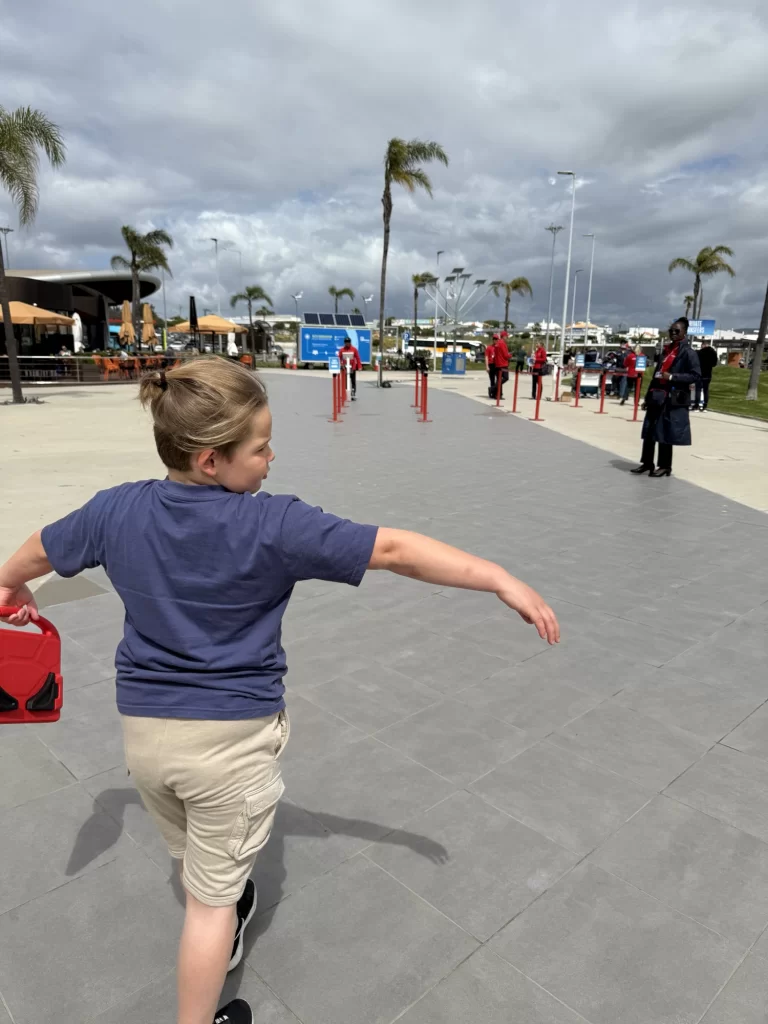 A boy in a blue shirt and beige shorts walks on a paved path, holding a red object, with palm trees and several people in the background under a cloudy sky.