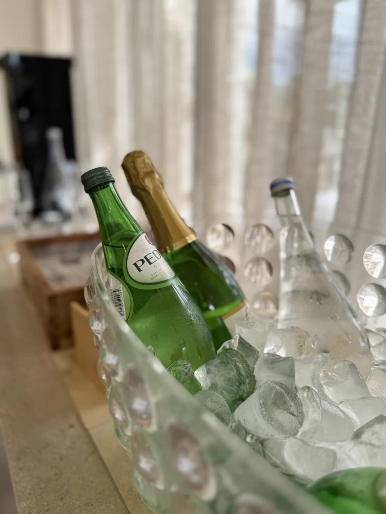 Three bottles, including sparkling water and champagne, are chilled in a glass ice bucket on a countertop, with a blurred background.