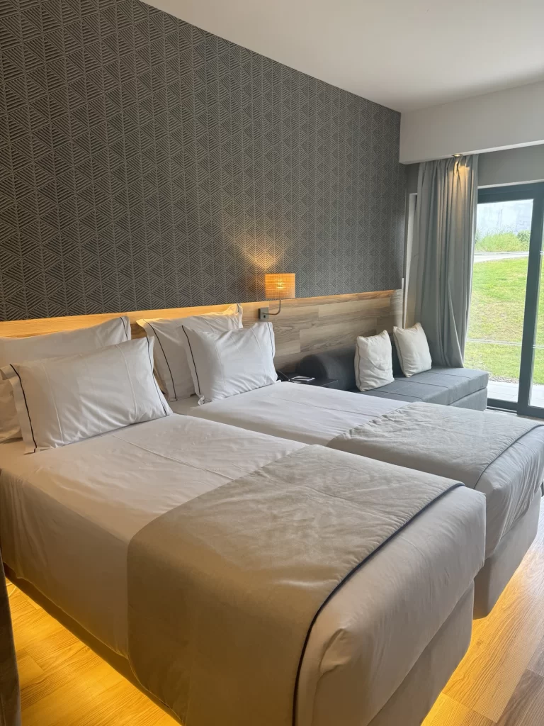 Modern hotel room with two single beds, a gray sofa, patterned accent wall, wood flooring, and a glass door leading to a grassy outdoor area.