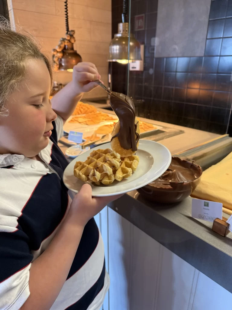 A child pours melted chocolate over a plate of waffles at a buffet station.
