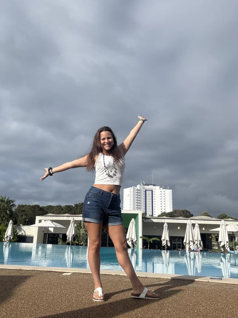 A woman stands with arms outstretched and smiling in front of a swimming pool, with closed umbrellas and a hotel building in the background under a cloudy sky.
