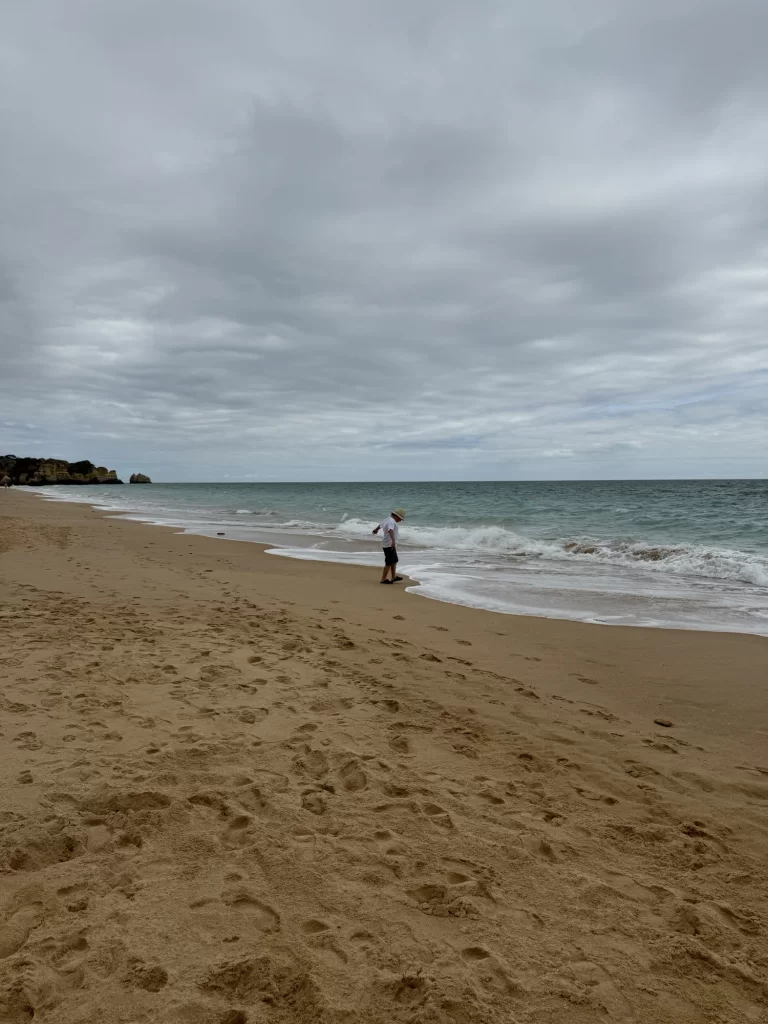 A person stands at the edge of the water on a sandy beach under a cloudy sky, with footprints visible in the sand.