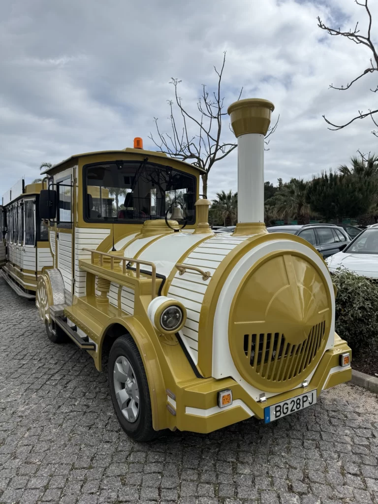 A yellow and white tourist road train is parked on a cobblestone surface near trees and cars under a cloudy sky.