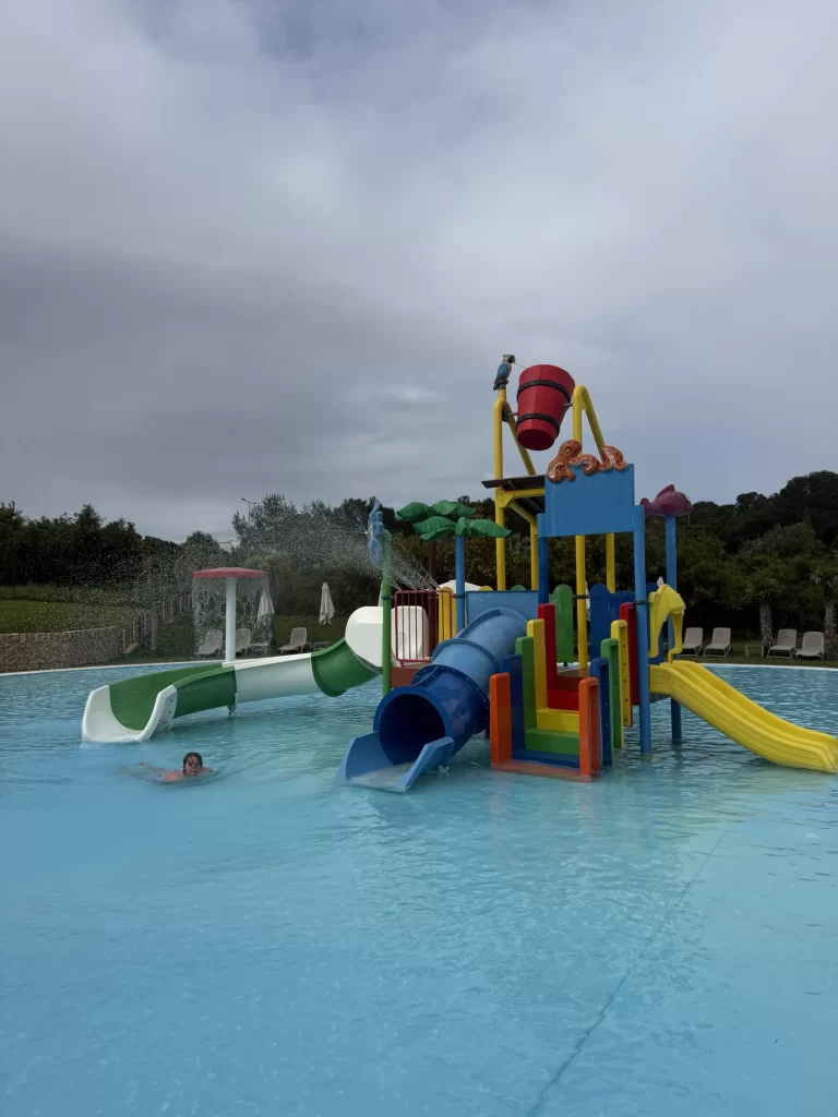 Colorful outdoor water play structure with slides, a tipping bucket, and stairs in a shallow pool; one child is swimming nearby under a cloudy sky.