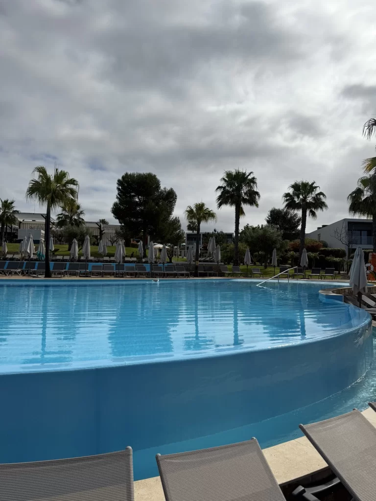 Circular outdoor swimming pool with clear blue water, surrounded by lounge chairs, palm trees, and closed umbrellas under a cloudy sky.