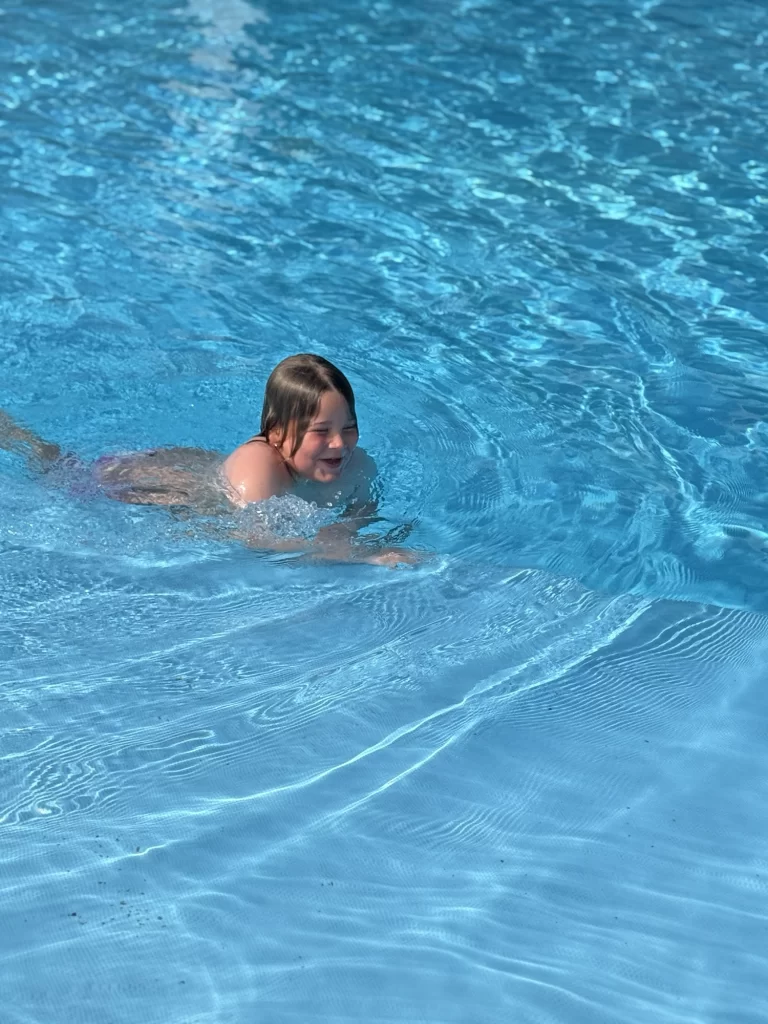 A young child swims in a clear blue outdoor pool, smiling and moving through the water on a sunny day.
