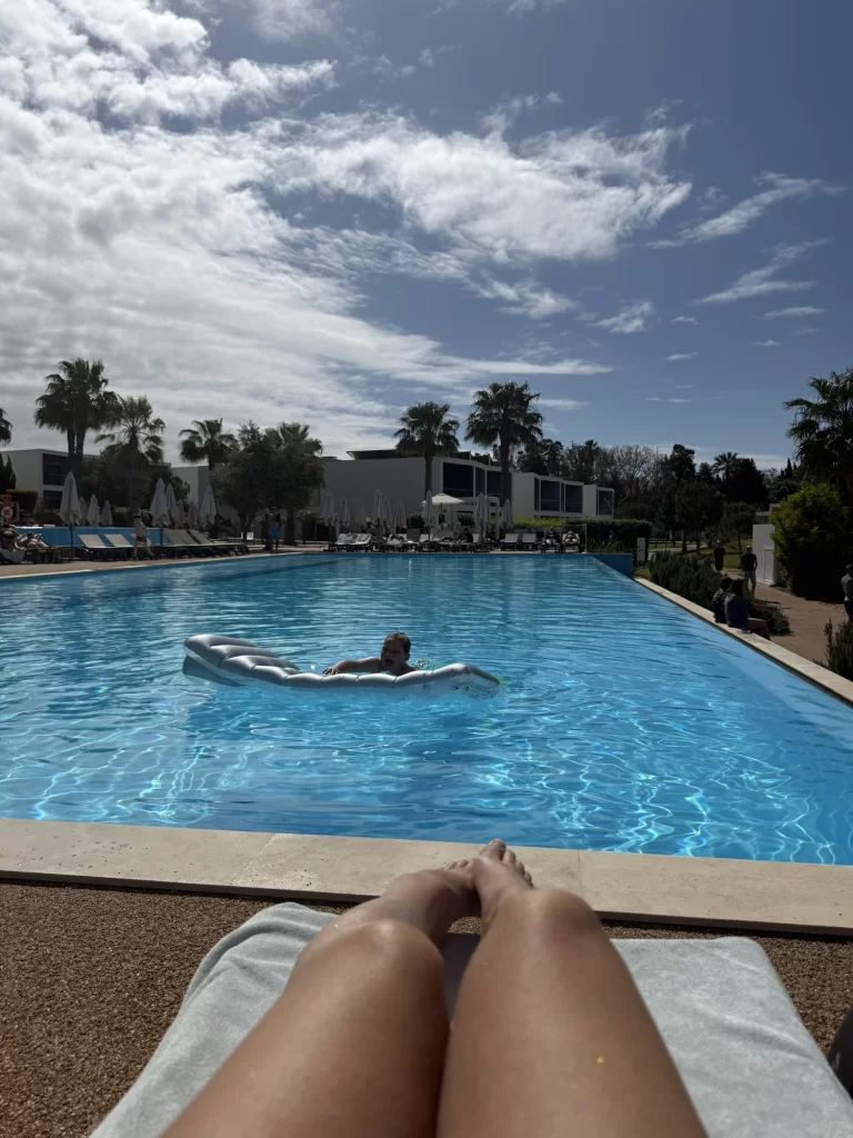 Person lying on a lounge chair by a pool with another person floating on an inflatable raft in the water; palm trees and buildings in the background under a partly cloudy sky.