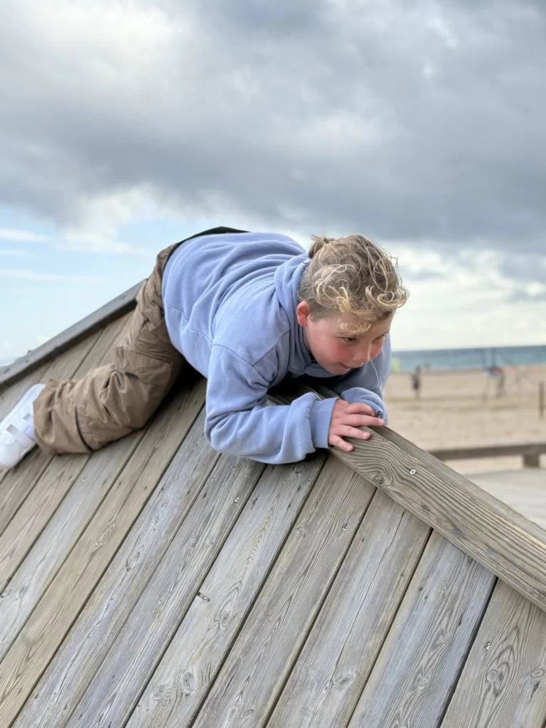A child in a blue hoodie and brown pants climbs a slanted wooden structure at a beach, with sand and ocean visible in the background.