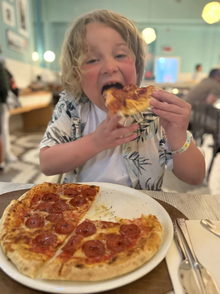 A young child with blond hair eats a slice of pepperoni pizza at a restaurant table, with the pizza on a plate in front of them.