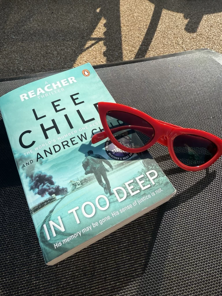 A copy of the book "In Too Deep" by Lee Child and Andrew Child beside red sunglasses on a textured dark surface in sunlight.