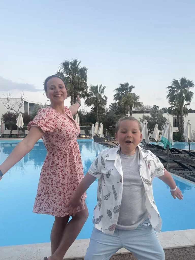 A woman in a pink floral dress and a child in a white shirt pose with arms outstretched in front of a swimming pool, with palm trees and lounge chairs in the background.