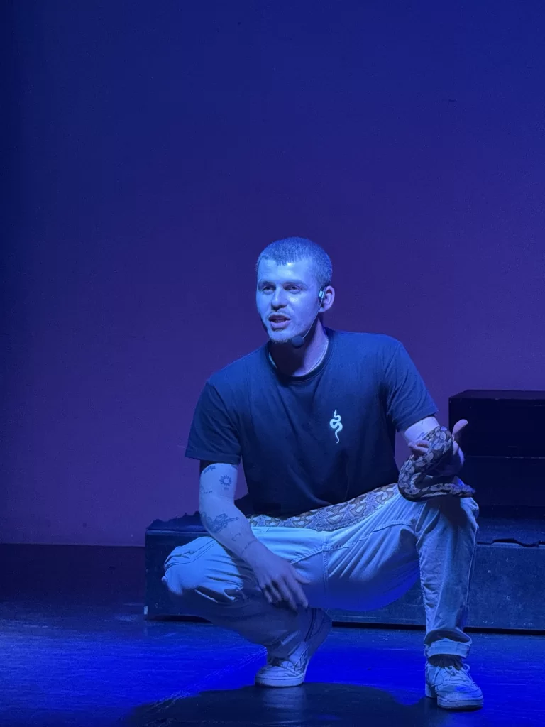 A person squats on stage under blue lighting, wearing a black t-shirt, light pants, and white sneakers, with visible tattoos and patterned cloth around their waist.