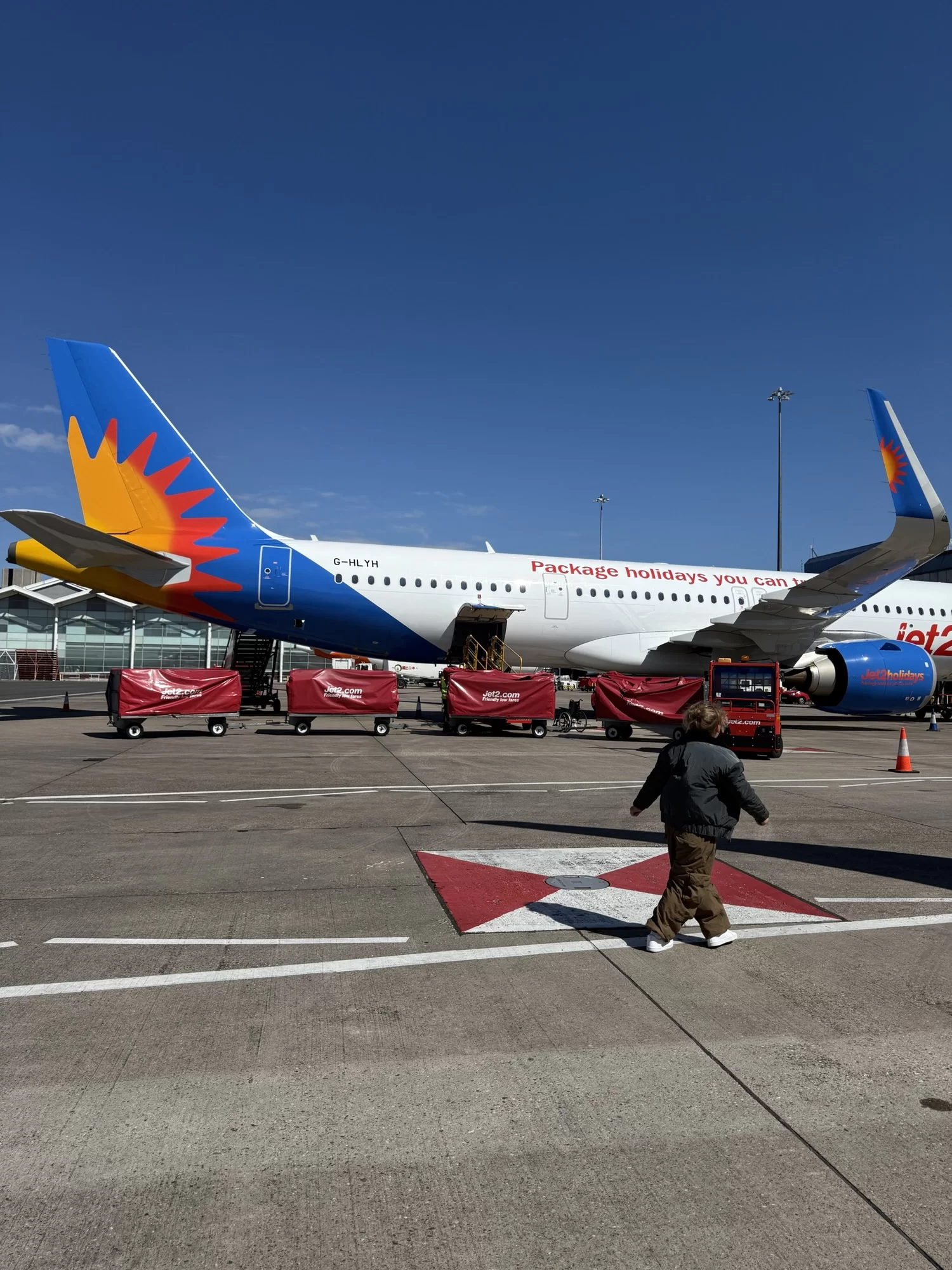 A passenger walks on the tarmac near a parked airplane with red baggage carts beside it under a clear blue sky.