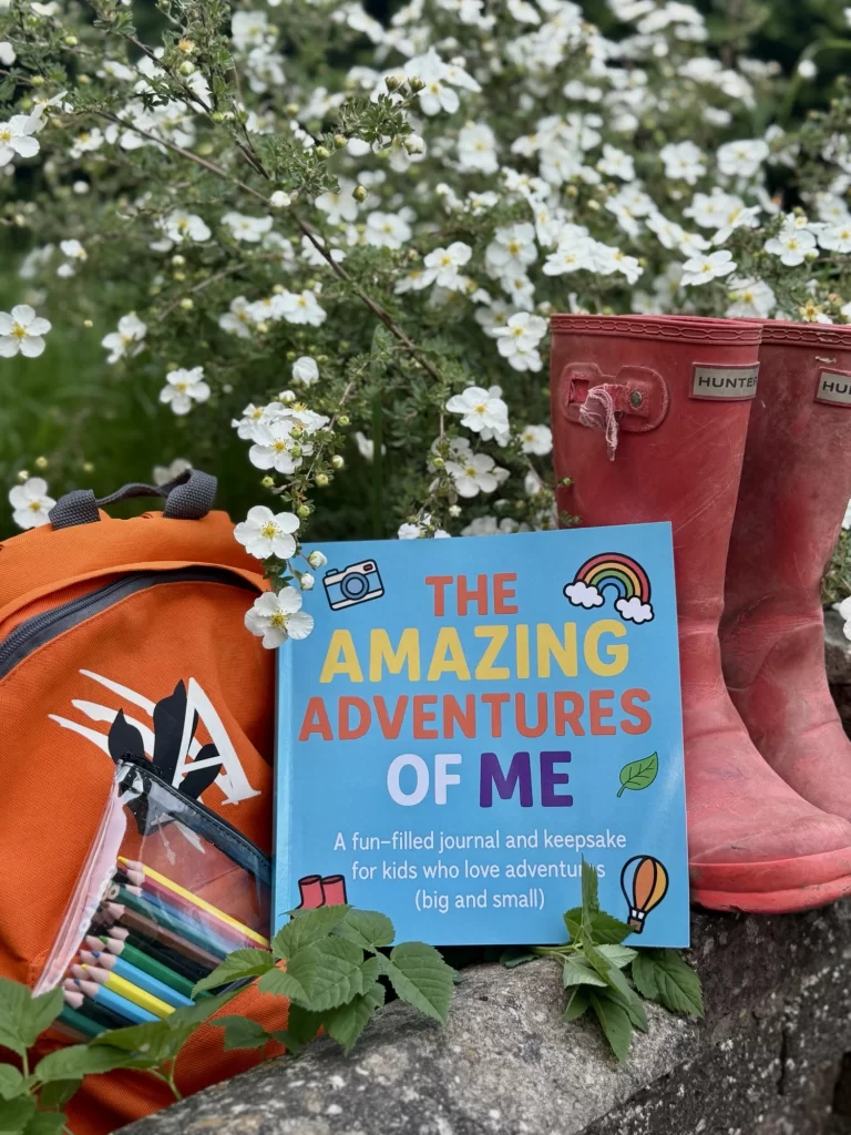 A children's adventure journal, red rubber boots, and an orange backpack with art supplies rest on a stone wall, with white flowers and greenery in the background.
