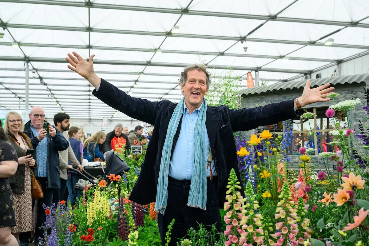 Man standing with arms outstretched among colorful flowers at an indoor garden show, with people observing in the background.