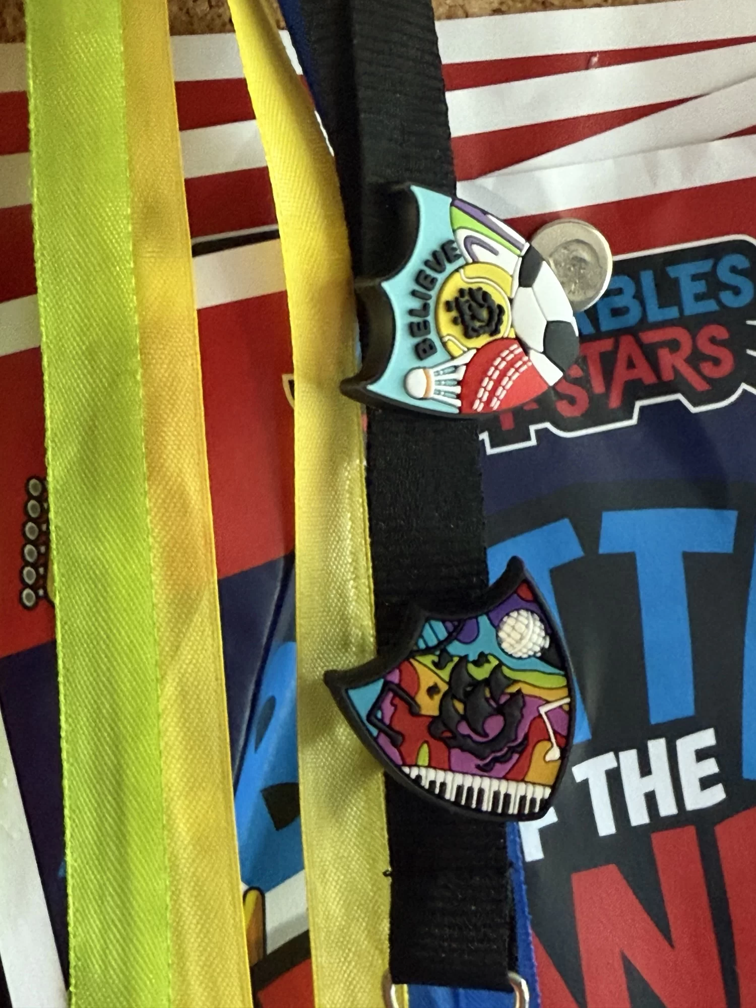 Two pins attached to black lanyards hang beside yellow ribbons; one pin features a soccer ball, the other shows a piano and colorful design. Printed papers are visible in the background.
