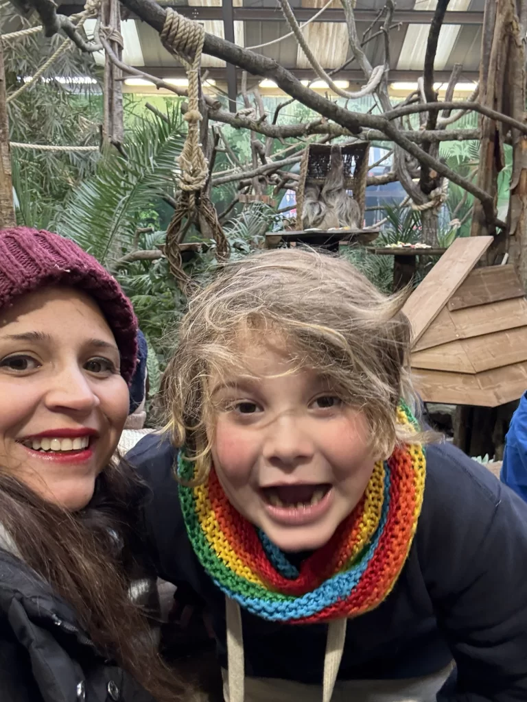 A woman and a child with a colorful scarf smile at the camera in an indoor zoo exhibit with branches and greenery in the background.