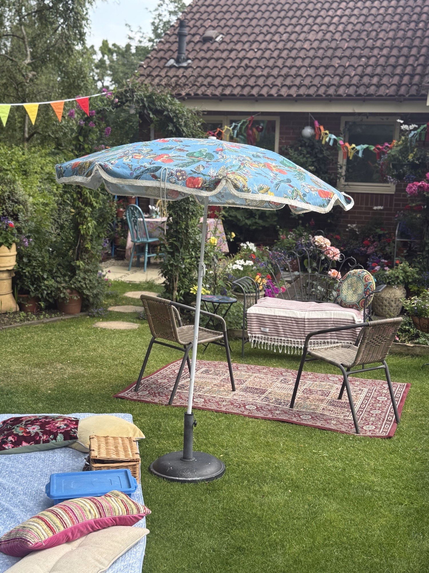 A back garden features a table and chairs shaded by a blue-patterned umbrella, with colourful bunting, rugs, and cushions set out on the grass near the house.