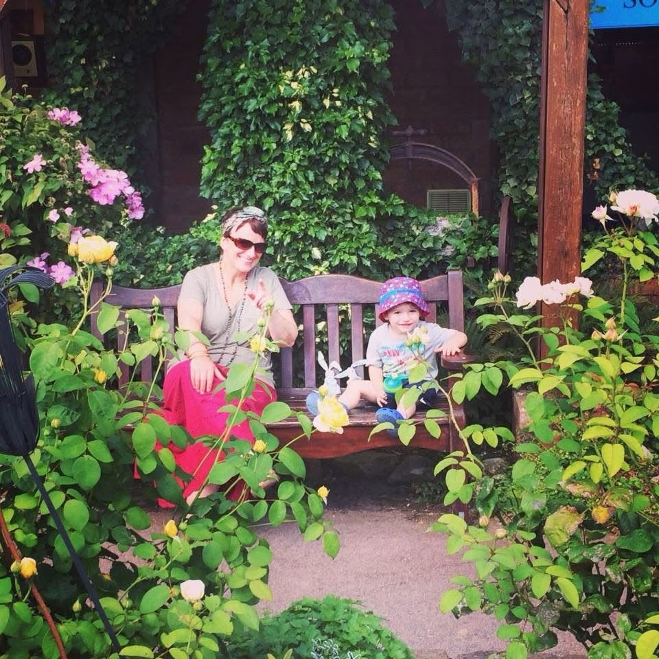 An adult and a child sit on a wooden bench in a garden surrounded by green plants and blooming flowers.