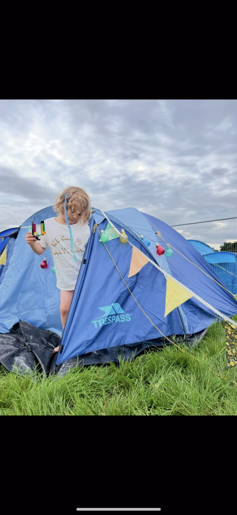 A child stands on grass in front of a blue tent decorated with bunting and fairy lights, beneath a cloudy sky.