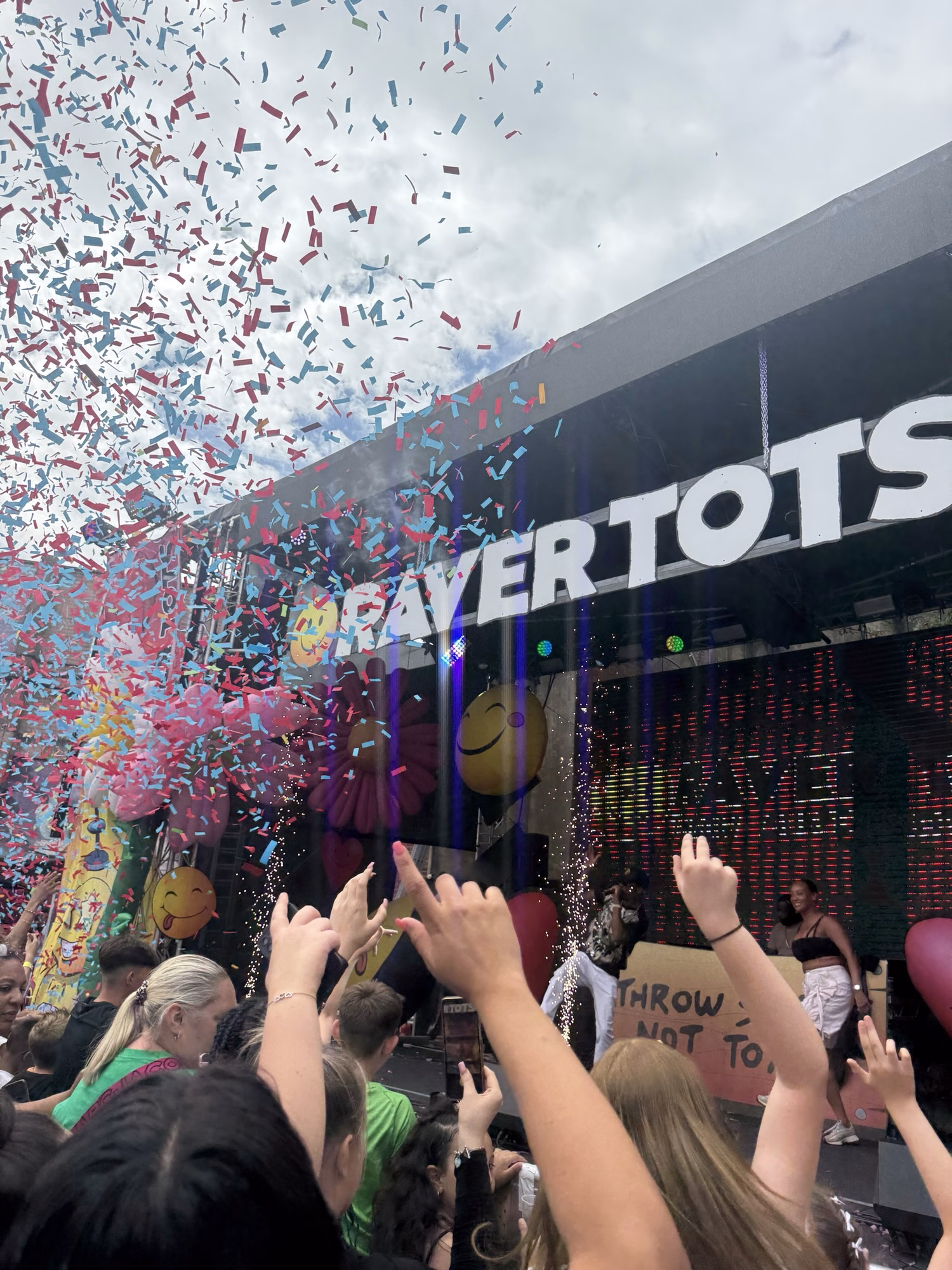 People at an outdoor concert cheer with raised hands as confetti falls and "RAVERTOTS" lights up above the performers on stage.