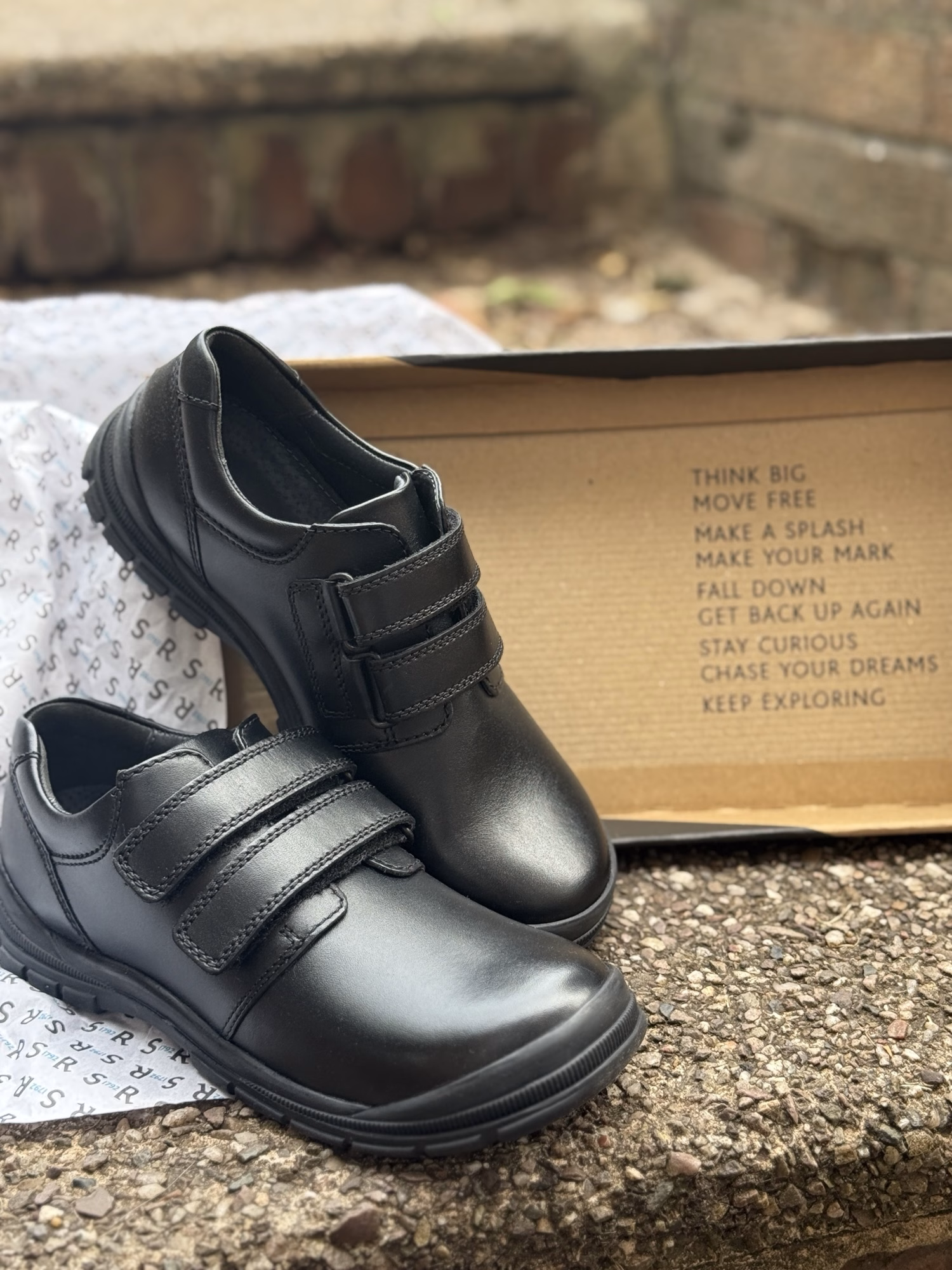 A pair of new black shoes with Velcro straps sits outside in front of an open shoebox, showing motivational phrases printed inside the lid.