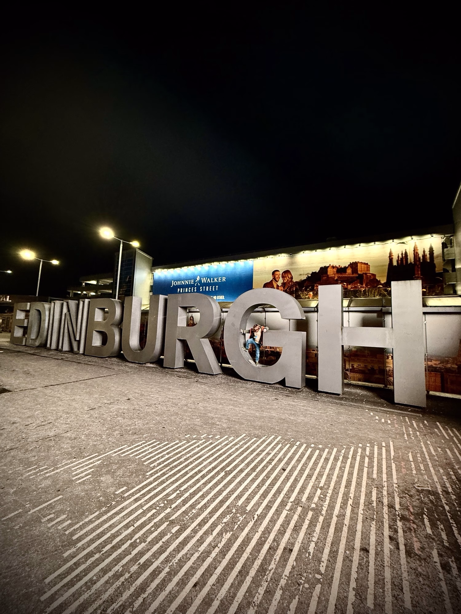 Silver letters spelling "EDINBURGH" stand outdoors at night, with a bright hoarding and dark sky behind them.