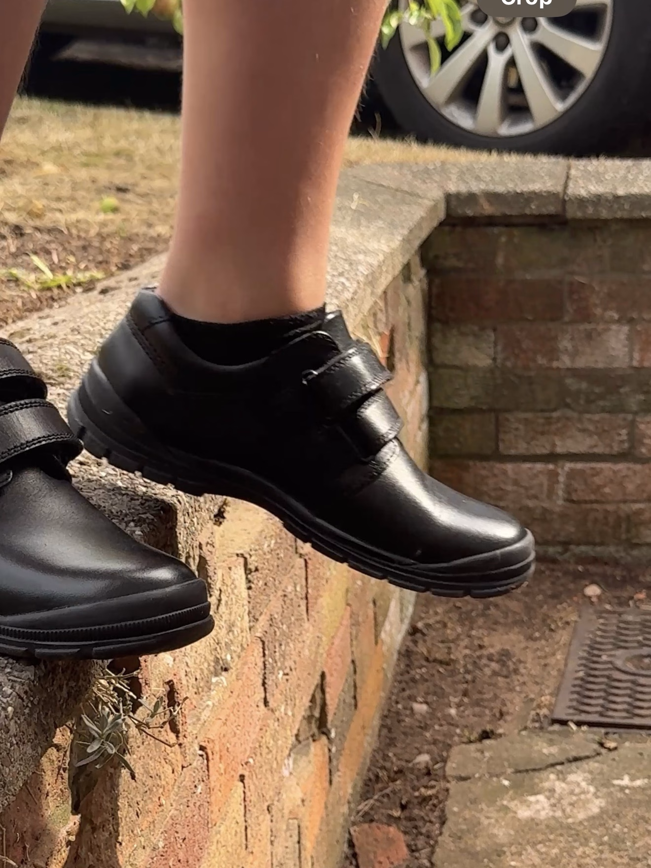 Someone in black Velcro shoes stands on the edge of a brick wall beside a pavement and a parked car.