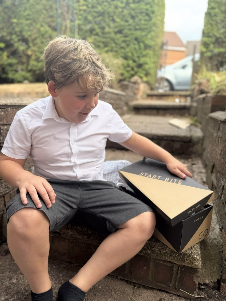 A young boy in a white shirt and grey shorts sits on outdoor steps, looking into a shoebox with the lid partly open.
