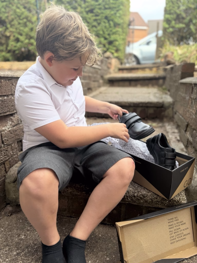A young boy sits on outdoor steps, wearing a white shirt and grey shorts, taking a black shoe from a box with another open box beside him.