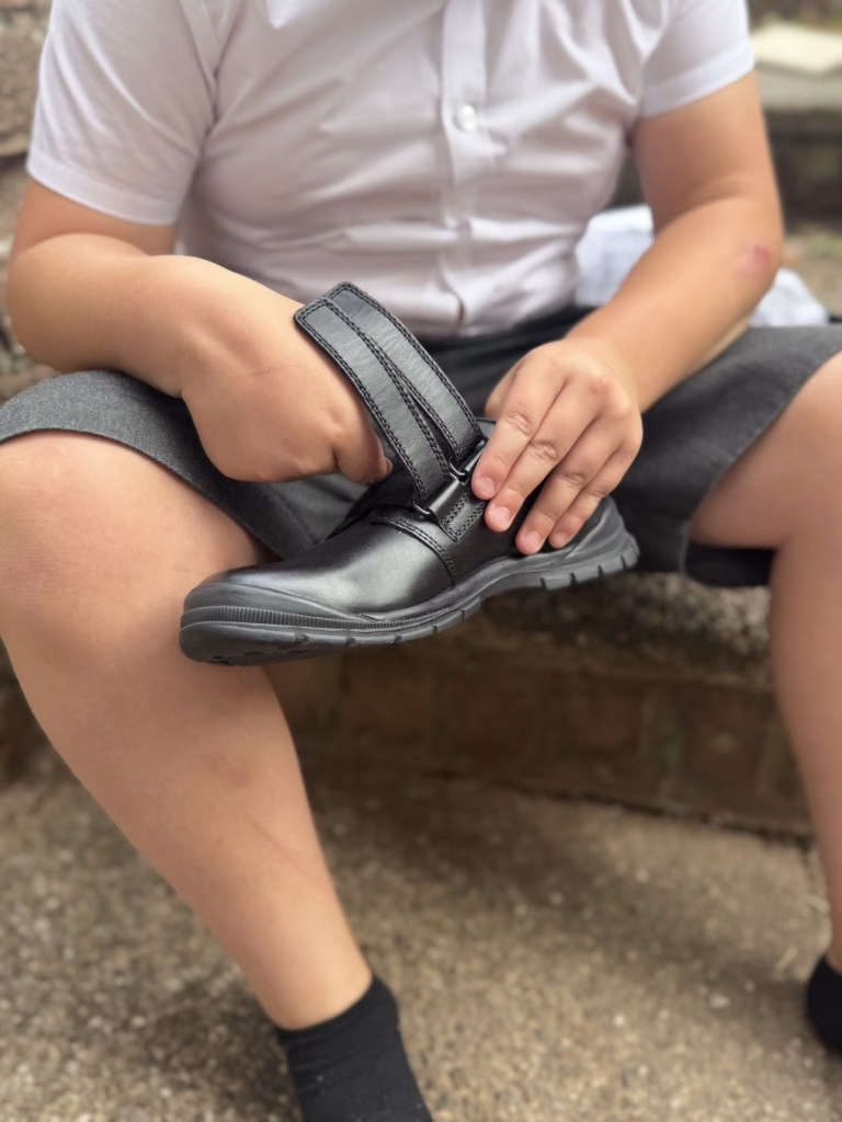 A child sits on outdoor steps, fastening the velcro strap on a black school shoe.