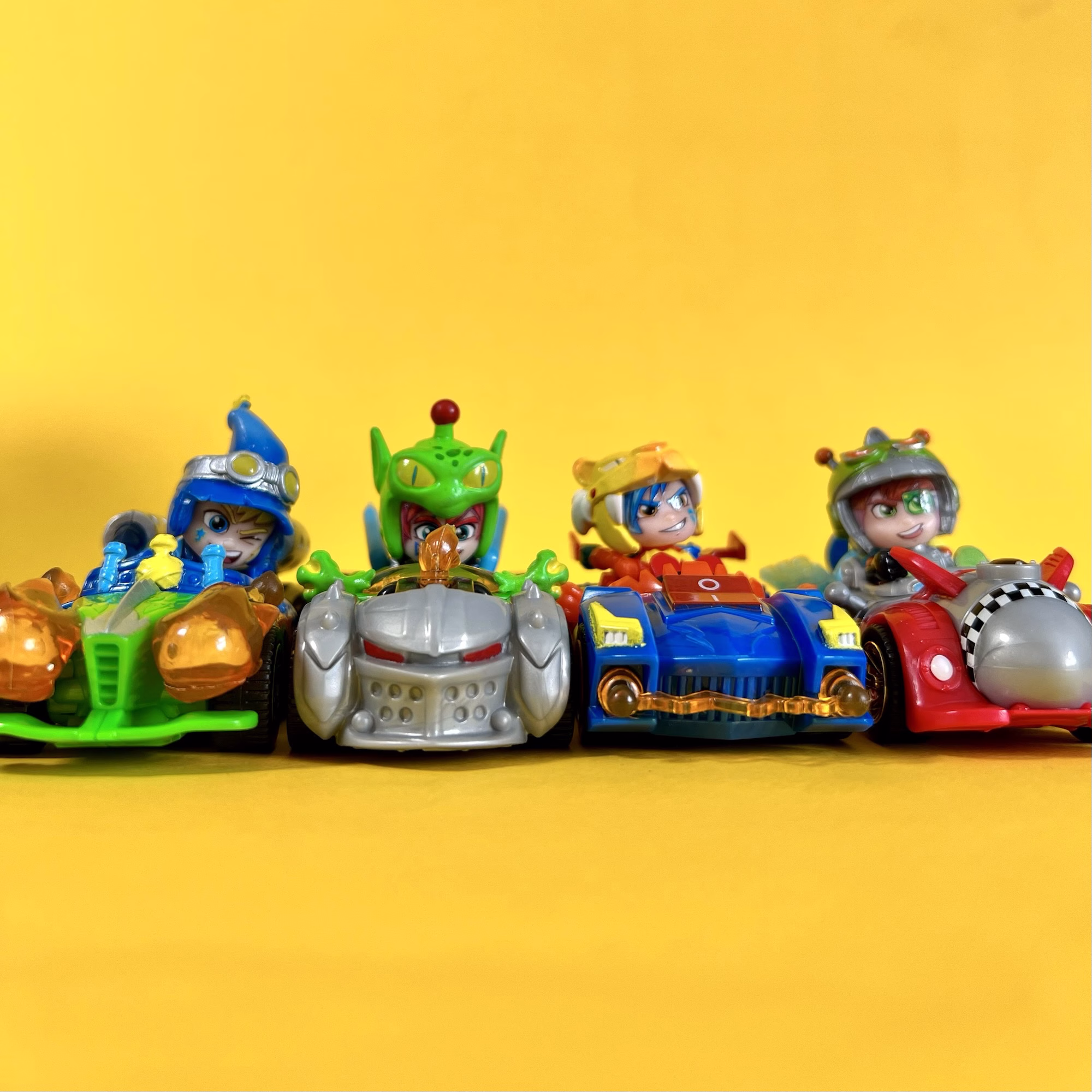 Four toy racing cars with cartoon drivers are lined up side by side on a yellow background.