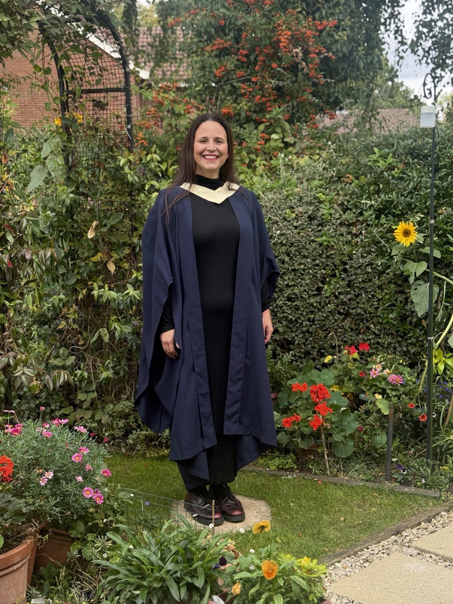 A smiling graduate in cap and gown stands in a flower-filled garden with lots of greenery.