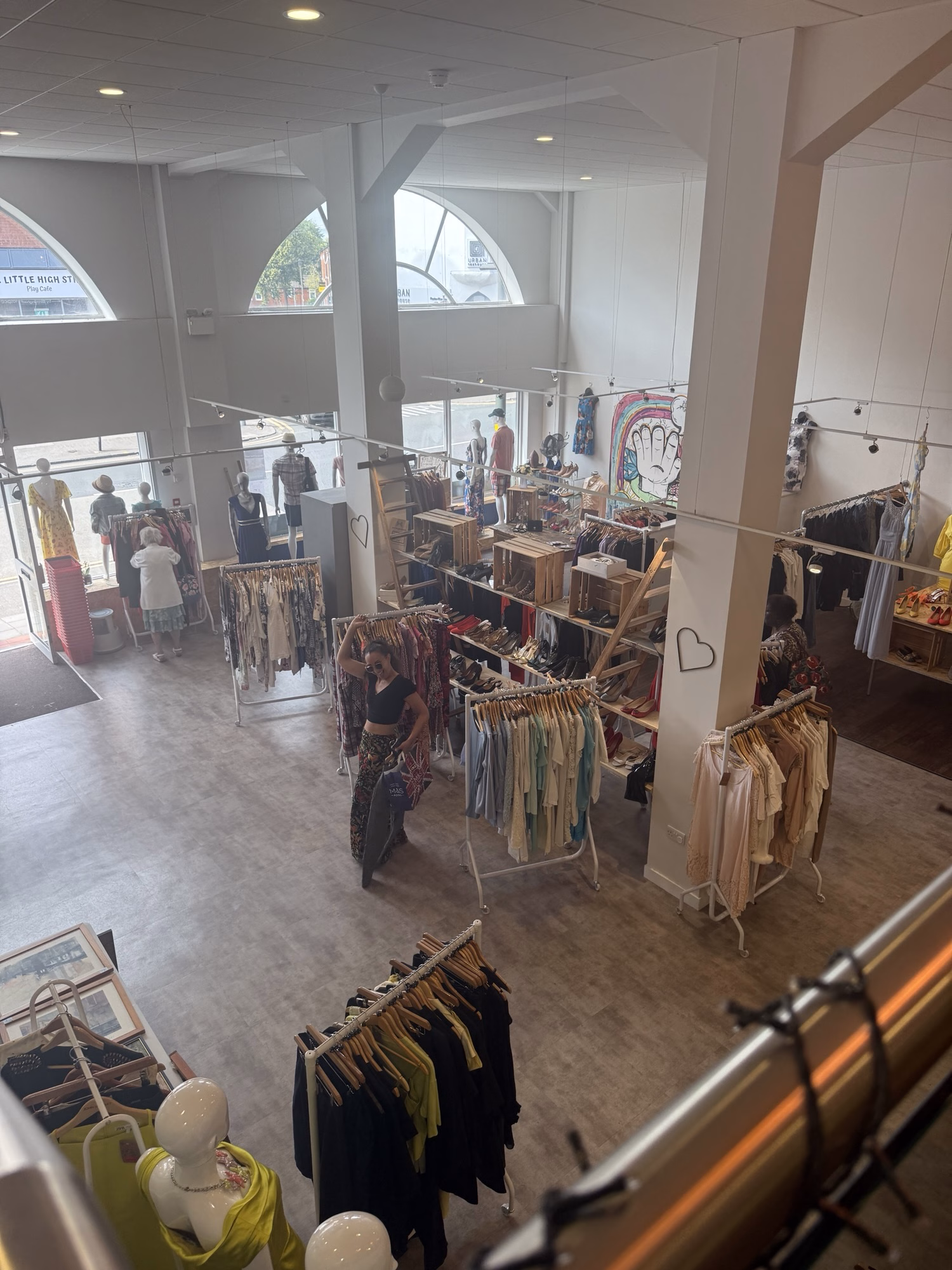 A clothes shop with large windows, shoppers browsing rails, and mannequins displayed at the front.