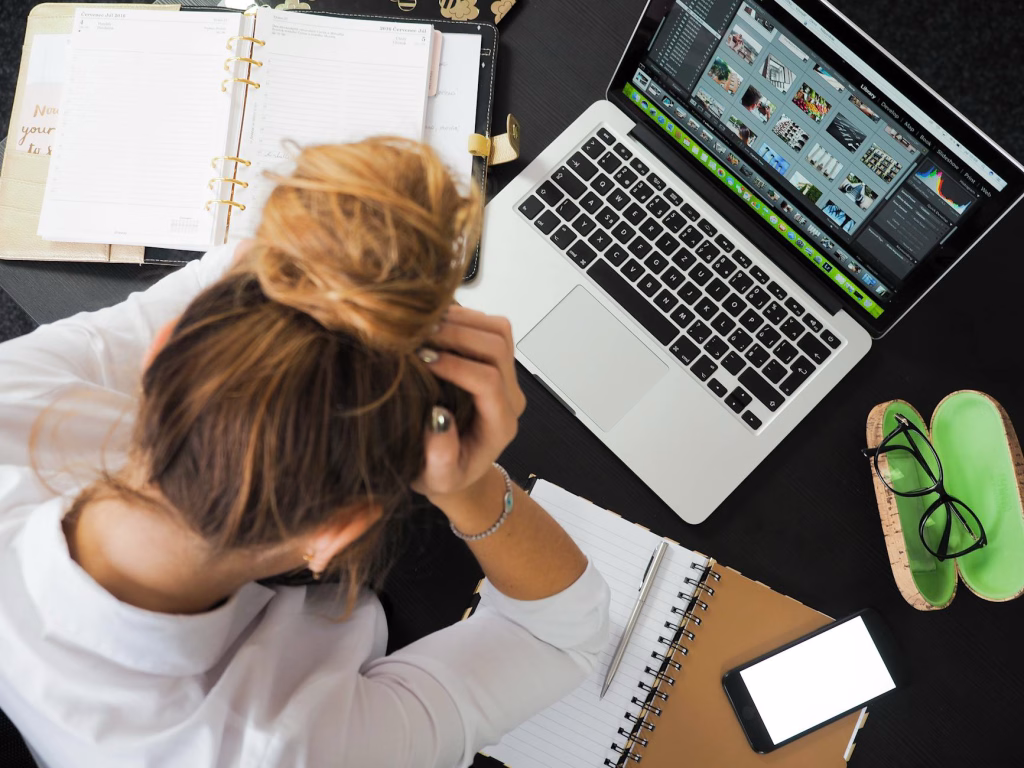 Someone sits at a desk with a laptop and papers, holding their head in their hands and looking stressed.